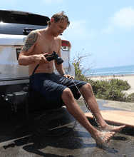 Load image into Gallery viewer,  Pictured is a male model sitting on a SMRT Tent Trailer Hitch Mount made of steel. He is wearing swim shorts/trunks and is using the spray nozzle hose of The Day Tank 3.85 Gallon to wash off his legs and feet. Perfect for your Canada roof top tent camping trip! Available for sale by SMRT Tent in Edmonton, Canada