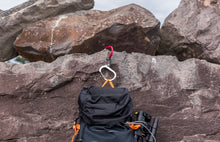 Load image into Gallery viewer, Pictured in metallic red for the open swivel hook and silver for the rest of the clip is a medium-sized metal carabiner hooked onto a rock ledge and holding aloft a black hiking backpack by its orange strap. This is a HeroClip Medium in Hot Rod Red. Perfect for your roof top tent adventure camp tool hanging needs! Available for sale in other colours (but not this colour) by SMRT Tent in Edmonton, Canada.