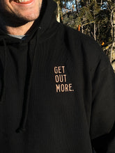 Load image into Gallery viewer, Pictured is a close-up view of the words Get Out More. in light Golden stitching on the upper-left chest of the male model's drawstring hoodie. The background is a coniferous tree. Chill out around your SMRT Tent roof top tent or get out more by hiking in this cozy hoodie! Available for sale by SMRT Tent in Edmonton, Canada.