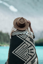 Load image into Gallery viewer, Pictured is a woman with her back to the camera, with a green & white mountain-patterned blanket wrapped around her. This is the Heartprint Threads 'Tracing the Columbia' Mountain Flakes Line Queen Blanket, created by Ecuadorian Artisans. Throw this warm blanket over your sleeping bag while camping outdoors! Available for sale at SMRT Tent in Edmonton, Canada.