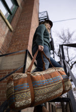 Load image into Gallery viewer, Pictured is a sky blue, terracotta orangey brown, grey, pale yellow, light brown and white patterned bag (similar looking to a duffle bag) with soft, brown leather straps. This is the Heartprint Threads 'Cambie' Weekend Traveller Bag, created by Ecuadorian Artisans. Perfect for all your week-long packing, storing and travelling needs! Available for sale at SMRT Tent in Edmonton, Canada.
