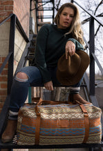 Load image into Gallery viewer, Pictured is a sky blue, terracotta orangey brown, grey, pale yellow, light brown and white patterned bag (similar looking to a duffle bag) with soft, brown leather straps lying on a stair step. This is the Heartprint Threads 'Cambie' Weekend Traveller Bag, created by Ecuadorian Artisans. Perfect for all your week-long packing, storing and travelling needs! Available for sale at SMRT Tent in Edmonton, Canada.