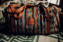 Load image into Gallery viewer, Pictured is a dark blue, light blue, black, terracotta brown, tan and brown patterned bag (similar looking to a duffle bag) with soft, brown leather straps. This is the Heartprint Threads 'Cartagena 2.0' Weekend Traveller Bag, created by Ecuadorian Artisans. Perfect for all your week-long packing, storing and travelling needs! Available for sale at SMRT Tent in Edmonton, Canada.