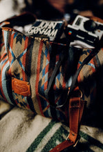 Load image into Gallery viewer, Pictured is a dark blue, light blue, black, terracotta brown, tan and brown patterned bag (similar looking to a duffle bag) with soft, brown leather straps. The bag’s zipper is open and showing its packed insides. This is the Heartprint Threads 'Cartagena 2.0' Weekend Traveller Bag, created by Ecuadorian Artisans. Perfect for all your week-long packing, storing and travelling needs! Available for sale at SMRT Tent in Edmonton, Canada.