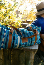 Load image into Gallery viewer, Pictured is a dark deep blue, dark ocean blue, bright sky blue, turquoise blue, teal and white patterned bag (similar looking to a duffle bag) with soft, brown leather straps This is the Heartprint Threads 'Galiano 2.0' Weekend Traveller Bag, created by Ecuadorian Artisans. Perfect for all your week-long packing, storing and travelling needs! Available for sale at SMRT Tent in Edmonton, Canada.