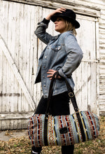 Load image into Gallery viewer, Pictured is a light blue, black, dark red, orangey-red, cream, dark brown and dark golden-yellowish tan patterned bag (similar looking to a duffle bag) with soft, black leather straps. This is the Heartprint Threads 'Hometown' Weekend Traveller Bag, created by Ecuadorian Artisans. Perfect for all your week-long packing, storing and travelling needs! Available for sale at SMRT Tent in Edmonton, Canada.
