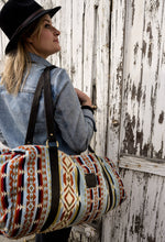 Load image into Gallery viewer, Pictured is a light blue, black, dark red, orangey-red, cream, dark brown and dark golden-yellowish tan patterned bag (similar looking to a duffle bag) with soft, black leather straps. This is the Heartprint Threads 'Hometown' Weekend Traveller Bag, created by Ecuadorian Artisans. Perfect for all your week-long packing, storing and travelling needs! Available for sale at SMRT Tent in Edmonton, Canada.