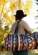 Load image into Gallery viewer, Pictured is a dark blue, muted aqua blue, dark wine red, blue, orange, pale green-ish yellow, cream and red patterned bag (similar looking to a duffle bag) with soft, black leather straps. This is the Heartprint Threads 'Magic' Weekend Traveller Bag, created by Ecuadorian Artisans. Perfect for all your week-long packing, storing and travelling needs! Available for sale at SMRT Tent in Edmonton, Canada.
