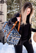 Load image into Gallery viewer, Pictured is a black, white, grey- slate blue- light blue, light grey and blue patterned bag (similar looking to a duffle bag) with soft, brown leather straps. This is the Heartprint Threads 'The Sunbreaker' Weekend Traveller Bag, created by Ecuadorian Artisans. Perfect for all your week-long packing, storing and travelling needs! Available for sale at SMRT Tent in Edmonton, Canada.
