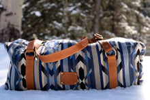 Load image into Gallery viewer, Pictured is a black, white, grey- slate blue- light blue, light grey and blue patterned bag (similar looking to a duffle bag) with soft, brown leather straps lying on snow. This is the Heartprint Threads 'The Sunbreaker' Weekend Traveller Bag, created by Ecuadorian Artisans. Perfect for all your week-long packing, storing and travelling needs! Available for sale at SMRT Tent in Edmonton, Canada.