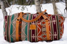 Load image into Gallery viewer, Pictured is a maroon, light tan, beige, brown, dark reddish brown, cream, teal, and orange patterned bag (similar looking to a duffle bag) with soft, orangey-brown leather straps lying on the snow. This is the Heartprint Threads 'Two Jack' Weekend Traveller Bag, created by Ecuadorian Artisans. Perfect for all your week-long packing, storing and travelling needs! Available for sale at SMRT Tent in Edmonton, Canada.