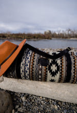 Load image into Gallery viewer, Pictured is a dark blue, grey, black, brown, greyish tan and white patterned bag (similar looking to a duffle bag) with soft, black leather straps and a brimmed, orange hat leaning on it. This is the Heartprint Threads 'Your City' Weekend Traveller Bag, created by Ecuadorian Artisans. Perfect for all your week-long packing, storing and travelling needs! Available for sale at SMRT Tent in Edmonton, Canada.