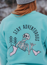 Load image into Gallery viewer, Pictured from the waist up, is our model with her back facing the camera. The back view shows our White laughing skeleton above and overlapping a mountain range with two simple pine trees to its right (our left) and the words Stay Adventurous written in Black above the image. 2 parts of the mountain range are shaded by a light pink. This SMRT Tent Crewneck in Teal is perfect to bring along on your overland roof top tent camping adventure! Available for sale by SMRT Tent in Edmonton, Canada.