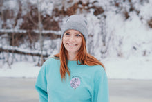 Load image into Gallery viewer, Pictured from the lower-chest up, is a ginger-haired, female model with fair skin and ocean blue eyes smiling and facing the camera. She is wearing the SMRT Tent Stay Adventurous Skeleton Crewneck in Teal (and a grey toque). The background is part of a frozen lake in a forested area. The surroundings are heavily covered in snow. This Crewneck is perfect to bring along on your overland roof top tent camping adventure! Available for sale by SMRT Tent in Edmonton, Canada.