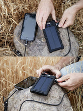 Load image into Gallery viewer, Pictured in two images above and below each other, is the portable, black Weatherproof Solar Power Bank with a black carabiner laying on a rock and connected by a USB cable to a smartphone and charging said phone. Perfect while camping or hiking for all of your recharging and flashlight needs! Available for sale by SMRT Tent in Edmonton, Canada.