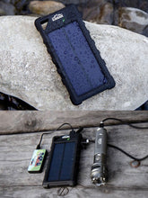 Load image into Gallery viewer, Pictured are two images above and below each other. In both is the portable, black Weatherproof Solar Power Bank. In the above image, the Power Bank is laying on a white rock and has countless water droplets splashed all over it. In the bottom image, the power bank lays on a wooden plank floor charging and connected by USB cables to a Kanan Outdoors Flashlight and a smaller, non-solar powered power bank. Available for sale by SMRT Tent in Edmonton, Canada.