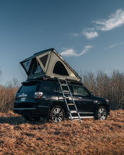 Load image into Gallery viewer, Pictured against a bright blue sky and plain brown grass, is a grey, completely set up hard shell roof top tent on the top of the roof of a black Toyota 4Runner SUV. This is the medium-sized Sky Loft, a 2-person hard shell roof top tent perfect for all overland roof top tent family outdoor camping! Available for sale by SMRT Tent in Edmonton, Canada.