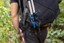 Load image into Gallery viewer, Pictured from their upper thighs to their upper arms is a hiker wearing a dark grey hiking backpack with their back facing the camera. In black is an adjustable rubber zip tie securely holding two pieces of gear to the bag. This is a zip tie from a Re-Ties Reusable Zip Ties 4-Pack. Perfect for your overland hiking gear managing, bundling, attaching, fastening and tying needs! Available for sale by SMRT Tent in Edmonton, Canada.