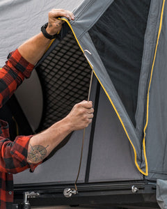 Pictured in the frame of the photo, from the left side, are a pair of hands (and arms) setting a replacement pole or strut to the tent flap of a fully unpacked hard shell roof top tent in Slate Grey. This is the perfect, large 2-person tent for any overland hard shell roof top tent camp adventure! Available for sale by SMRT Tent in Edmonton, Canada.