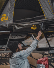 Load image into Gallery viewer, Pictured from the chest up with her back facing the camera, is a female model wearing sunglasses, a black toque, and an acid-washed jean jacket. She is reaching up and unzipping the packed-up SMRT Tent Camp Shower Kit. Her surroundings are a Toyota Tundra truck and her Summit Suite SMRT Tent Roof Top Tent in Grey. Perfect for your Canada roof top tent truck adventure! Available for sale by SMRT Tent in Edmonton, Canada.
