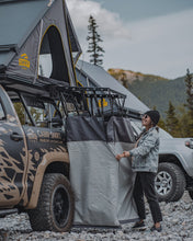 Load image into Gallery viewer, Pictured in full frame, is our female model in black jeans and her acid-washed jean jacket. She has fully set up the now unpacked SMRT Tent Camp Shower Kit. Her surroundings are a Toyota Tundra truck and her Summit Suite SMRT Tent Roof Top Tent in Grey. Perfect for your Canada roof top tent truck adventure! Available for sale by SMRT Tent in Edmonton, Canada.
