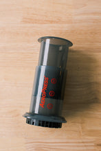 Load image into Gallery viewer, Pictured is a clear, grey cylinder, the name AeroPress is written lengthwise in red on its side. This is the portable Original AeroPress Coffee Press. Perfect for your overland roof top tent coffee, espresso and cold brew maker needs! Available for sale by SMRT Tent in Edmonton, Canada.
