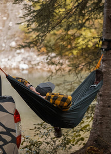 Pictured is our male model with his back to the camera as he lays in a hammock tied to a tree and his truck. He is wearing our SMRT Dark Grey snapback hat forwards and with the angle we only see the top and the bill of the hat. Keep the sun out of your eyes during your overland roof top tent camping adventure in this sweet hat! Available for sale by SMRT Tent in Edmonton, Canada.