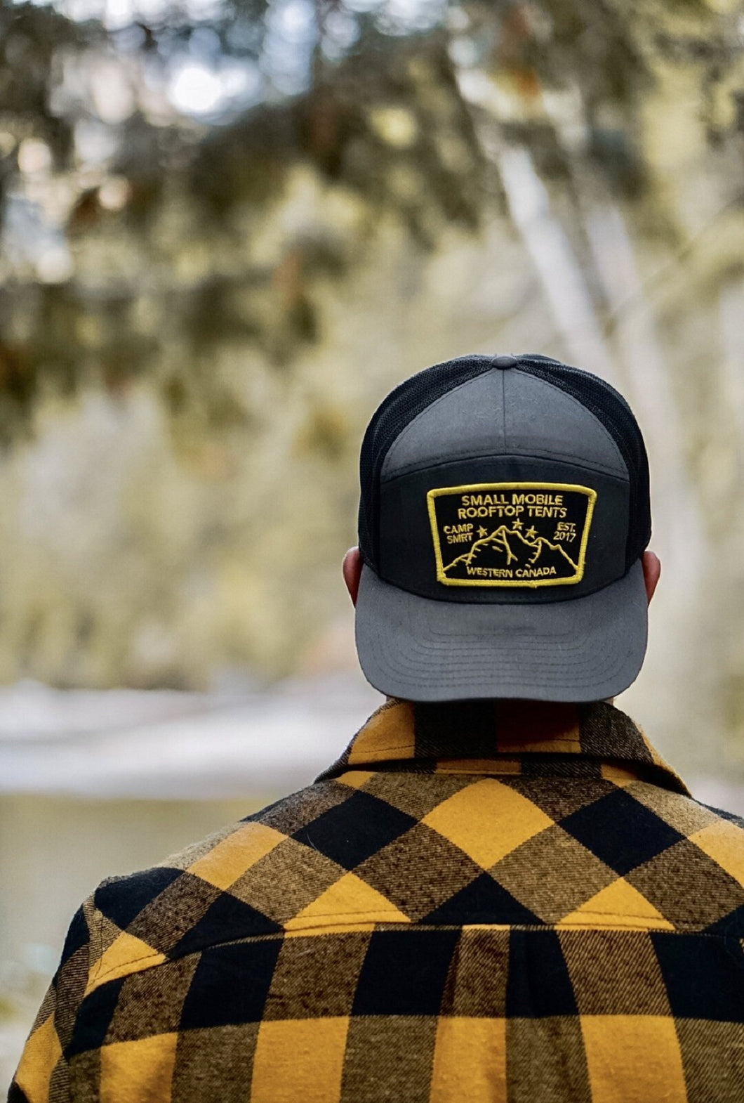 Pictured from the upper back to the head, is a male model with his back to the camera. He is wearing our SMRT Tent Dark Grey snapback hat backwards so that its front and patch face the camera. Keep the sun out of your eyes during your overland roof top tent camping adventure in this sweet hat!  Available for sale by SMRT Tent in Edmonton, Canada.