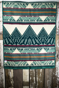 Pictured is a teal, dark turquoise green, orange, pomegranate red, burgundy and white coloured, mountain patterned baby blanket. This is the Heartprint Threads 'Little Adventurer' Baby Blanket, created by Ecuadorian Artisans. Perfect for your everyday soft & warm baby blanket needs! Available for sale at SMRT Tent in Edmonton, Canada.