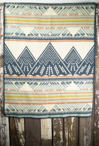 Pictured is a light blue, orange, golden yellow, light grey, dark slate blue and cream coloured, mountain patterned baby blanket. This is the Heartprint Threads 'Mountain Lion' Baby Blanket, created by Ecuadorian Artisans. Perfect for your everyday soft & warm baby blanket needs! Available for sale at SMRT Tent in Edmonton, Canada.