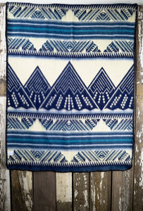 Pictured is a light slate blue, darkish aqua blue, light grey, pale sky blue, dark blue and white coloured, mountain patterned baby blanket. This is the unfolded Heartprint Threads 'Small and Mighty' Baby Blanket, created by Ecuadorian Artisans. Perfect for your everyday soft & warm baby blanket needs! Available for sale at SMRT Tent in Edmonton, Canada.