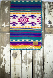 Pictured is a violet, lightly sandy white, bright yellow, pale yellow, bright orange, magenta, cream, purple and white coloured, traditional Aztec patterned baby blanket. This is the technicolour Heartprint Threads 'Wild One' Baby Blanket, created by Ecuadorian Artisans. Perfect for your everyday soft & warm baby blanket needs! Available for sale at SMRT Tent in Edmonton, Canada.