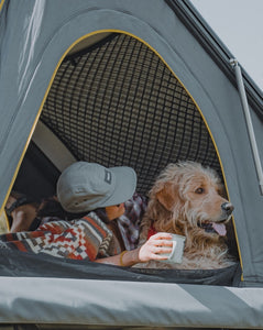 Pictured is a close-up of a SMRT Tent hard shell roof top tent in grey where a girl and her dog are next to a red, grey & olive, wave-patterned blanket. This is a Heartprint Threads ‘Mosaic Pebble’ Maskuy Collection Queen Blanket, created by Ecuadorian Artisans. Throw this warm blanket over your sleeping bag while camping outdoors! Available for sale at SMRT Tent in Edmonton, Canada.