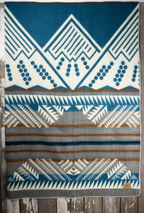 Pictured is a grey, brown, cream & Crayola cerulean blue, mountain-patterned and laid-out blanket. This is the Heartprint Threads 'Sky Blue Sky' Mountain Flakes Line Queen Blanket, created by Ecuadorian Artisans. Throw this warm blanket over your sleeping bag while camping outdoors! Available for sale at SMRT Tent in Edmonton, Canada.
