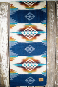 Pictured is a blue, patterned and laid-out blanket. This is the Heartprint Threads 'Teal Rustic' Queen Blanket, created by Ecuadorian Artisans. Throw this warm blanket over your sleeping bag while camping outdoors! Available for sale at SMRT Tent in Edmonton, Canada.
