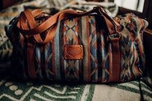 Load image into Gallery viewer, Pictured is a dark blue, light blue, black, terracotta brown, tan and brown patterned bag (similar looking to a duffle bag) with soft, brown leather straps. This is the Heartprint Threads &#39;Cartagena 2.0&#39; Weekend Traveller Bag, created by Ecuadorian Artisans. Perfect for all your week-long packing, storing and travelling needs! Available for sale at SMRT Tent in Edmonton, Canada.
