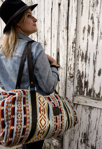 Pictured is a light blue, black, dark red, orangey-red, cream, dark brown and dark golden-yellowish tan patterned bag (similar looking to a duffle bag) with soft, black leather straps. This is the Heartprint Threads 'Hometown' Weekend Traveller Bag, created by Ecuadorian Artisans. Perfect for all your week-long packing, storing and travelling needs! Available for sale at SMRT Tent in Edmonton, Canada.