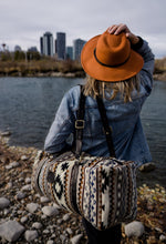 Load image into Gallery viewer, Pictured is a dark blue, grey, black, brown, greyish tan and white patterned bag (similar looking to a duffle bag) with soft, black leather straps being carried by a woman with her back to the camera. She is looking at part of the cityscape of Calgary. This is the Heartprint Threads &#39;Your City&#39; Weekend Traveller Bag, created by Ecuadorian Artisans. Perfect for all your week-long packing, storing and travelling needs! Available for sale at SMRT Tent in Edmonton, Canada.
