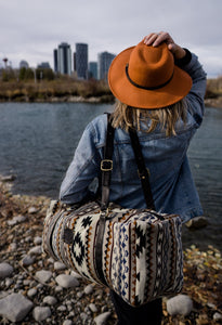 Pictured is a dark blue, grey, black, brown, greyish tan and white patterned bag (similar looking to a duffle bag) with soft, black leather straps being carried by a woman with her back to the camera. She is looking at part of the cityscape of Calgary. This is the Heartprint Threads 'Your City' Weekend Traveller Bag, created by Ecuadorian Artisans. Perfect for all your week-long packing, storing and travelling needs! Available for sale at SMRT Tent in Edmonton, Canada.