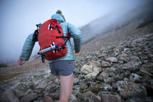 Load image into Gallery viewer, Pictured almost fully in frame with their back to the camera, is a hiker wearing a bright, baby blue puffy jacket and a bright red backpack. They have securely attached their LifeStraw to the two outside, lower hoop-straps of their backpack. The LifeStraw can be used to drink directly from lakes and streams and delivers clean and safe water to the drinker&#39;s mouth. Perfect for your overland roof top tent camp survival needs! Available for sale by SMRT Tent in Edmonton, Canada.
