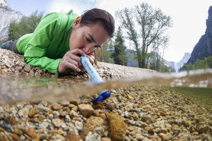 Pictured almost fully in frame and facing slightly away from the camera, is a brunette model wearing a bright, grass green jacket and laying beside a body of water. She is holding - into the water - in her hands and drinking from Lake Close with a LifeStraw. The LifeStraw can be used to drink directly from lakes and streams and delivers clean and safe water to the drinker's mouth. Perfect for your overland roof top tent camp survival needs! Available for sale by SMRT Tent in Edmonton, Canada.