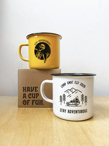 Pictured are the two variants of the SMRT Tent enamel mug. One is the Stay Adventurous Skeleton mug in yellow, set atop a brown cardboard box that says Have A Cup Of Fun. On the table and slightly to the right is the Camp SMRT EST 2020 Stay Adventurous in white. Available for purchase by SMRT Tent in Edmonton, Canada.