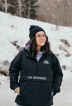 Load image into Gallery viewer, Pictured from the hips up, is a brunette, female model wearing glasses and a black toque looking just off-camera. She is wearing our SMRT Tent Rainy Jacket (in Black) with her hands inside the two sides of the front multi-pocket. Layer around your SMRT Tent roof top tent camp or SUV in this sweet anorak! Available for sale by SMRT Tent in Edmonton, Canada.
