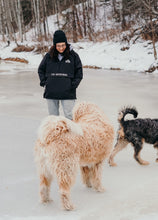 Load image into Gallery viewer, Pictured in the unfocused foreground are two large and fluffy dogs, making our brunette model visible only from the knees up in the focused mid-to-background. She is wearing our SMRT Tent Rainy Jacket (in Black) with her hands on the sides of the front multi-pocket. Layer around your SMRT Tent roof top tent camp or SUV in this sweet anorak! Available for sale by SMRT Tent in Edmonton, Canada.
