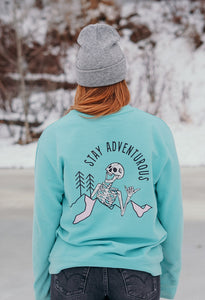 Pictured from the hips up, is our model with her back facing the camera. The back view shows our White laughing skeleton above and overlapping a mountain range with two simple pine trees to its right (our left) and the words Stay Adventurous written in Black above the image. 2 parts of the mountain range are shaded by a light pink, mimicking snow or daylight. This Crewneck is available for sale by SMRT Tent in Edmonton, Canada.