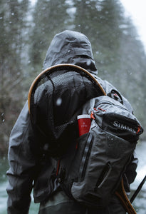 Pictured with their back facing the camera, is a hiker wearing a grey winter jacket with the hood up and a grey, single-strap SIMMS backpack on their right shoulder. It presses a large mesh net (the shape of which looks like an overly large tennis racket) to the model's back. A red VSSL First Aid is tucked into the side pocket of the backpack. Perfect for your hiking & ice-fishing first aid kit needs! Available for sale by SMRT Tent in Edmonton, Canada.