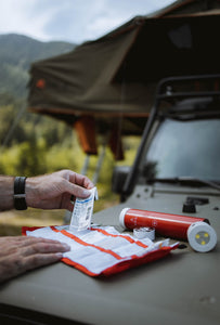 Pictured on the hood of a car is a VSSL First Aid cylinder, flashlight end cap unscrewed, while in front and slanted to the left of it is an unrolled red multi-pocket strip. From the left side of the frame, a pair of hands: the right laying on the unrolled med kit and car hood and the left hand pulling out an item from the roll. In-between the multi-pocket strip and the cylinder is a 20-foot roll of Adventure Tape. Perfect for any of your first aid needs! Available for sale by SMRT Tent in Edmonton, Canada.