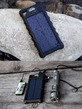 Load image into Gallery viewer, Pictured are two images above and below each other. In both is the portable, black Weatherproof Solar Power Bank. In the above image, the Power Bank is laying on a white rock and has countless water droplets splashed all over it. In the bottom image, the power bank lays on a wooden plank floor charging and connected by USB cables to a Kanan Outdoors Flashlight and a smaller, non-solar powered power bank. Available for sale by SMRT Tent in Edmonton, Canada.
