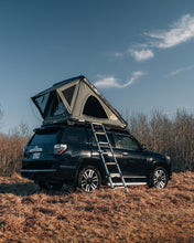 Load image into Gallery viewer, Pictured  against a bright blue sky and plain brown grass, is a grey, completely set up hard shell roof top tent on the top of the roof of a black Toyota 4Runner SUV. This is the medium-sized Sky Loft, a 2-person hard shell roof top tent perfect for all overland roof top tent family outdoor camping! Available for sale by SMRT Tent in Edmonton, Canada.

